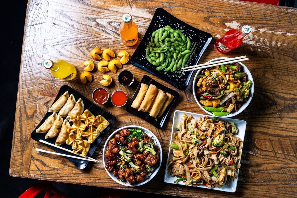 Overhead shot of various Chinese takeout dishes: dumplings, noodles, vegetables, spring rolls, edamame, and drinks on a wooden table.