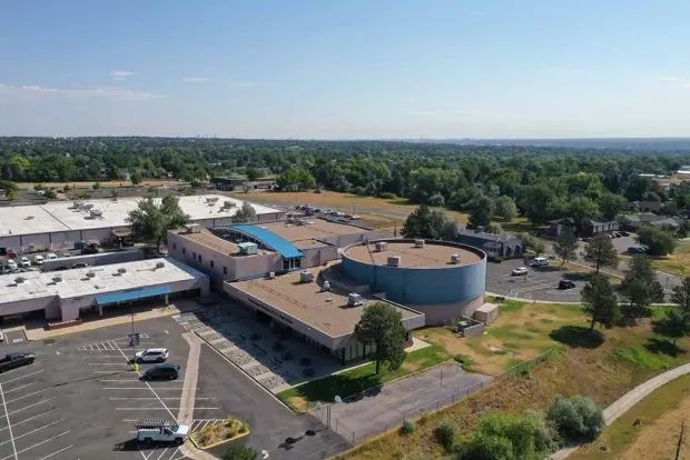 Aerial view of a light blue and beige building with a flat roof, surrounded by trees and a parking lot.