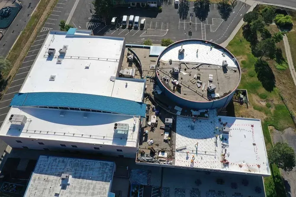 Aerial view of a complex with flat roofs, including a large cylindrical structure and a parking area.