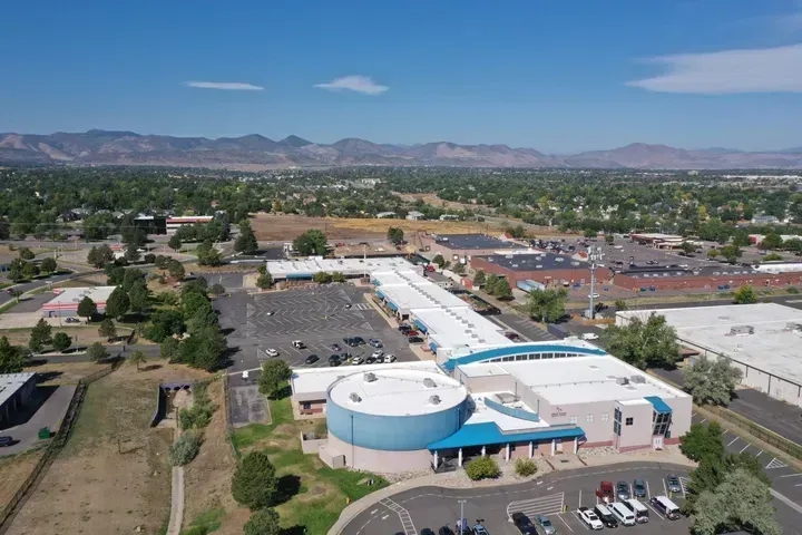 Aerial view of a large blue and white building complex with a parking lot and mountain backdrop on a sunny day.