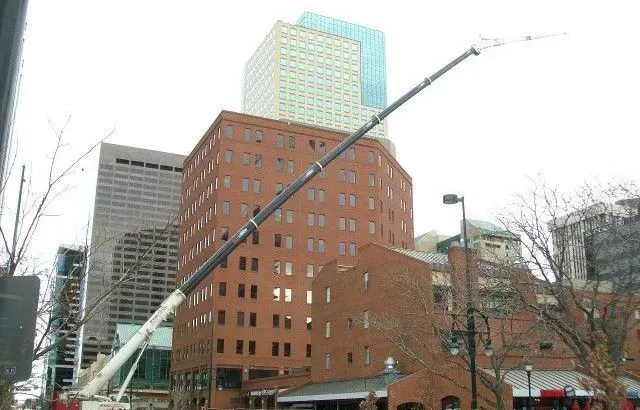 A tall crane extends over a brick building in a city, with other buildings in the background.