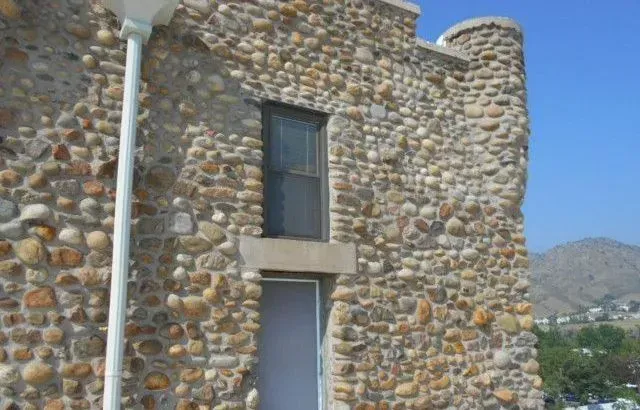 Stone-walled building with a window, a white door, and a circular tower on the right, against a blue sky.