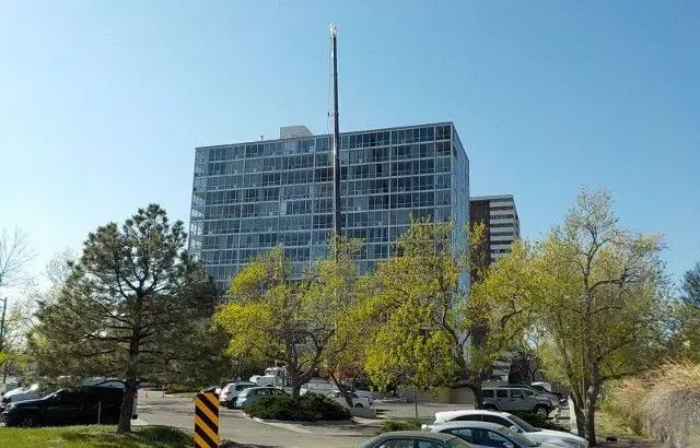 Tall glass building with antenna. Trees and cars in a parking lot on a sunny day.