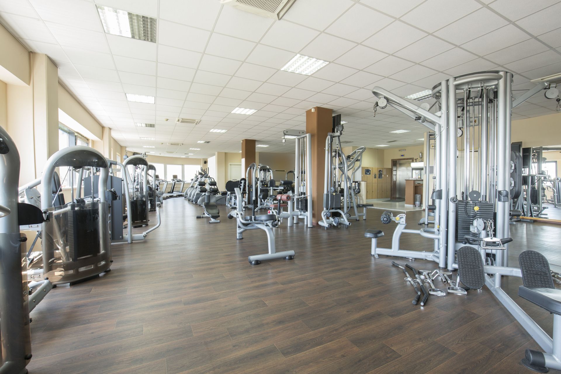 Bright gym with rows of exercise machines and wooden flooring under large ceiling lights