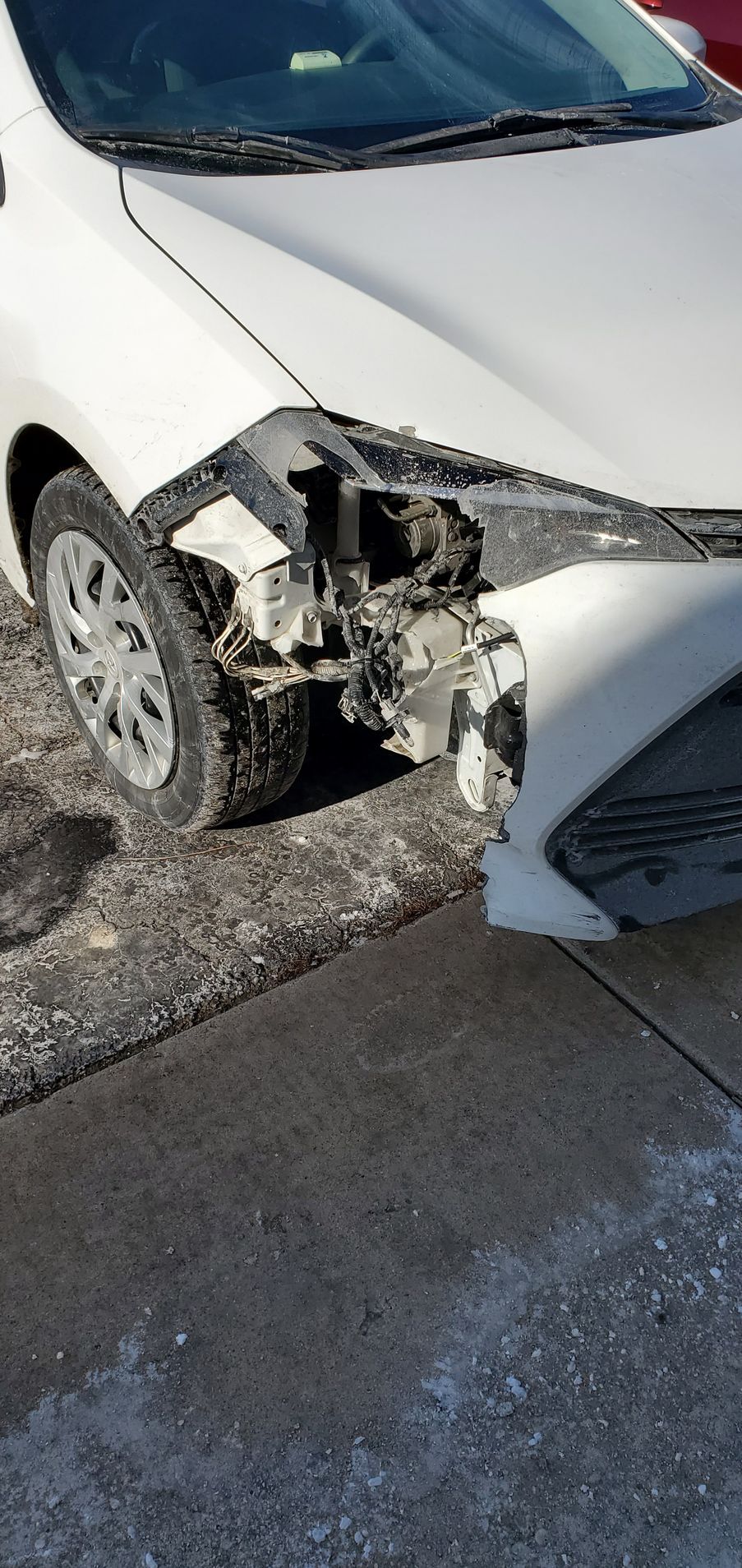 A white car with a damaged front bumper is parked on the side of the road.