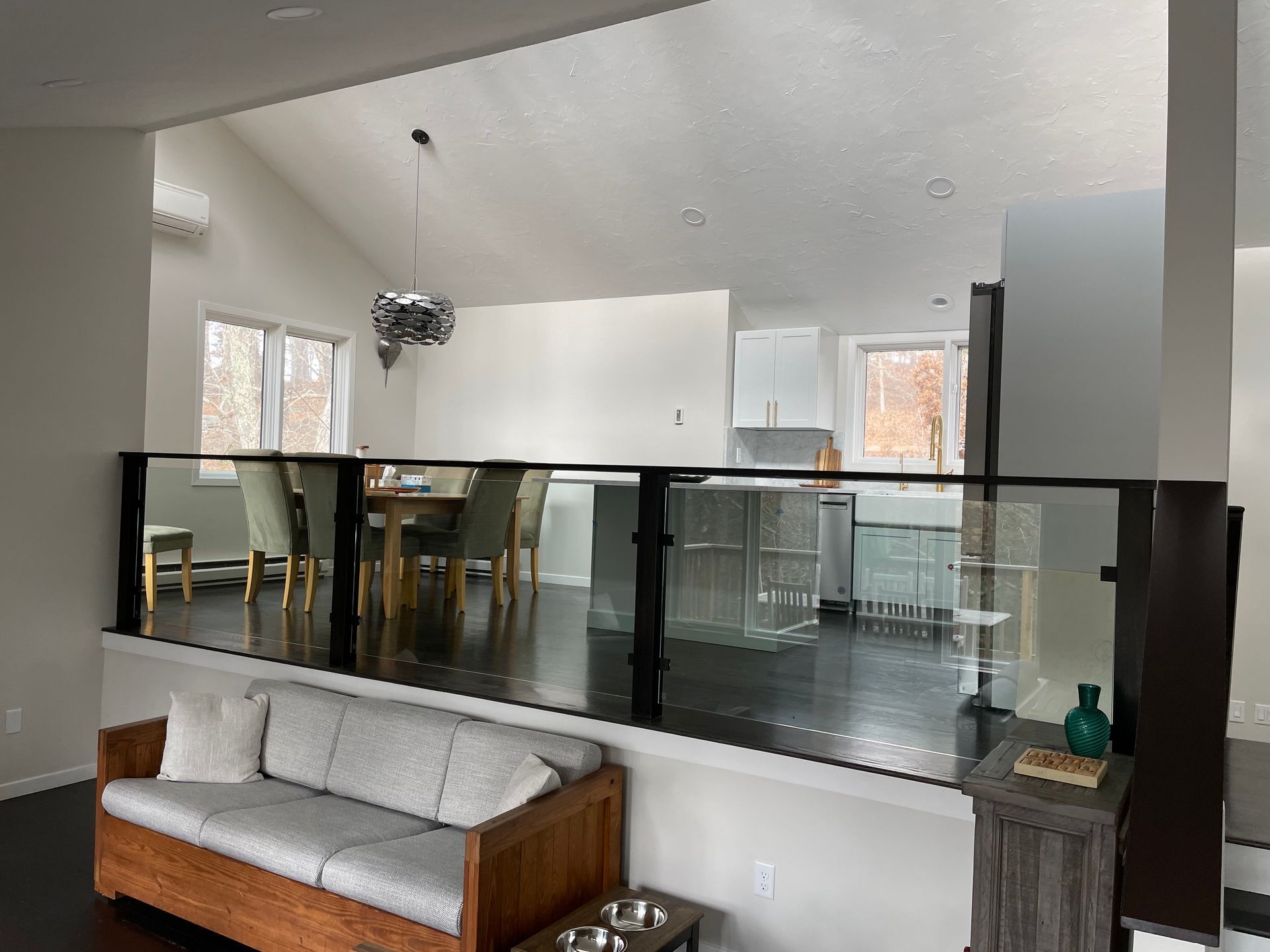 A living room with a sofa and a glass railing overlooking a kitchen and dining area with bright white walls and dark wood floors.