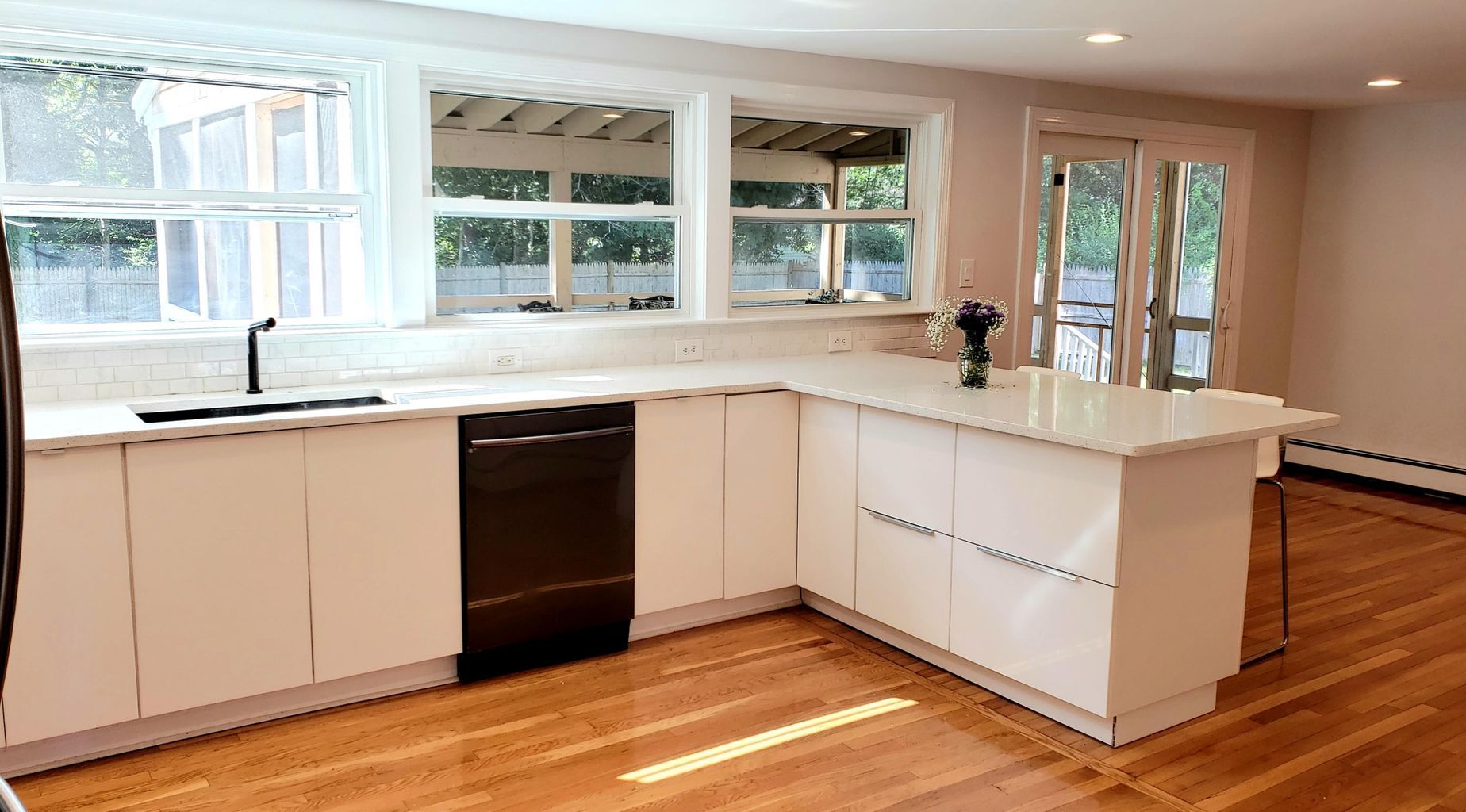 Modern kitchen with white cabinets, a dark dishwasher, and a marble countertop. Sunlight streams through the windows.