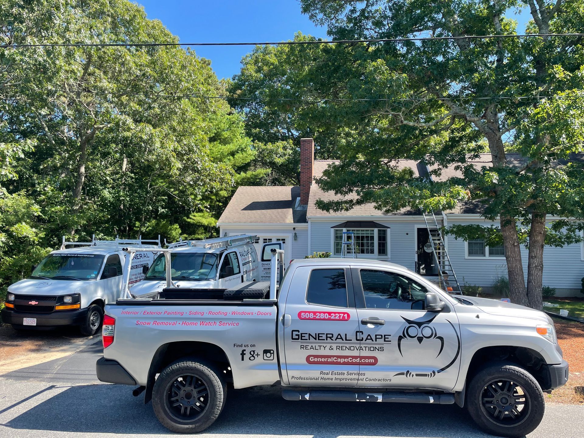 A residential property with three work vehicles parked in front. The vehicles have company logos that are visible. The sky is blue and sunny.