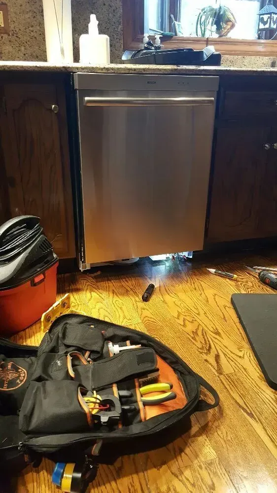A stainless steel dishwasher installed between wooden cabinets. Repair tools are on the floor in front of it.