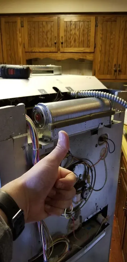 A person giving a thumbs-up near a machine in a kitchen, with wood cabinets in the background.