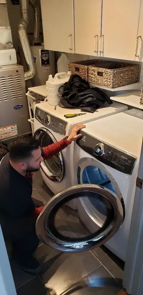 A man repairing a washing machine in a laundry room; holding the door open.