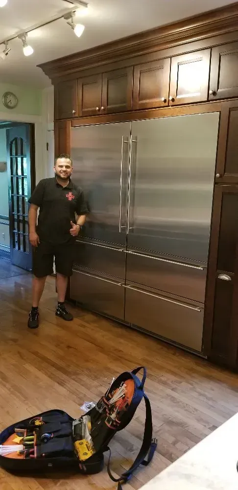 A man stands beside a large stainless steel refrigerator in a kitchen. A toolbox sits on the floor.