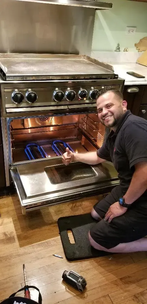 Man giving thumbs up, kneeling by an open oven. Oven interior glowing, kitchen setting.