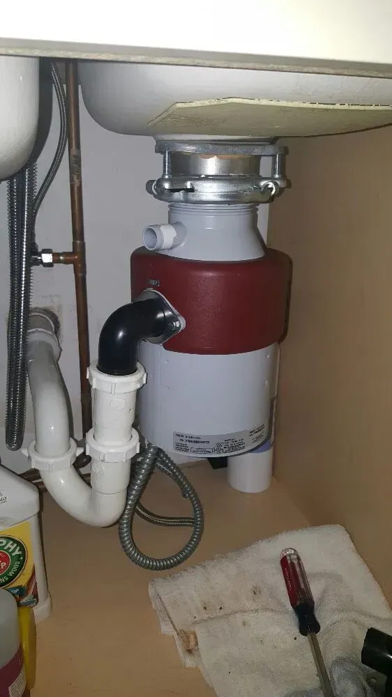 Garbage disposal under a kitchen sink with pipes and plumbing, in a cabinet, along with a screwdriver and rag.