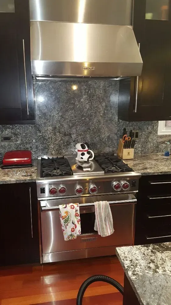 Stainless steel oven and range hood in a kitchen with dark cabinets and granite backsplash.