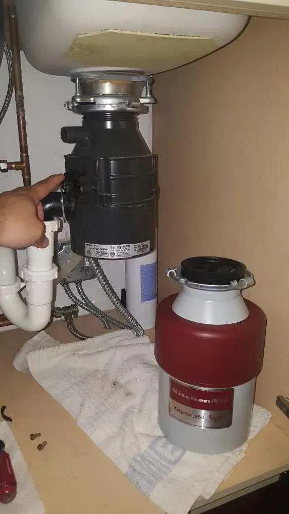 A person points to a black garbage disposal under a sink, next to a red and silver new one on a counter.