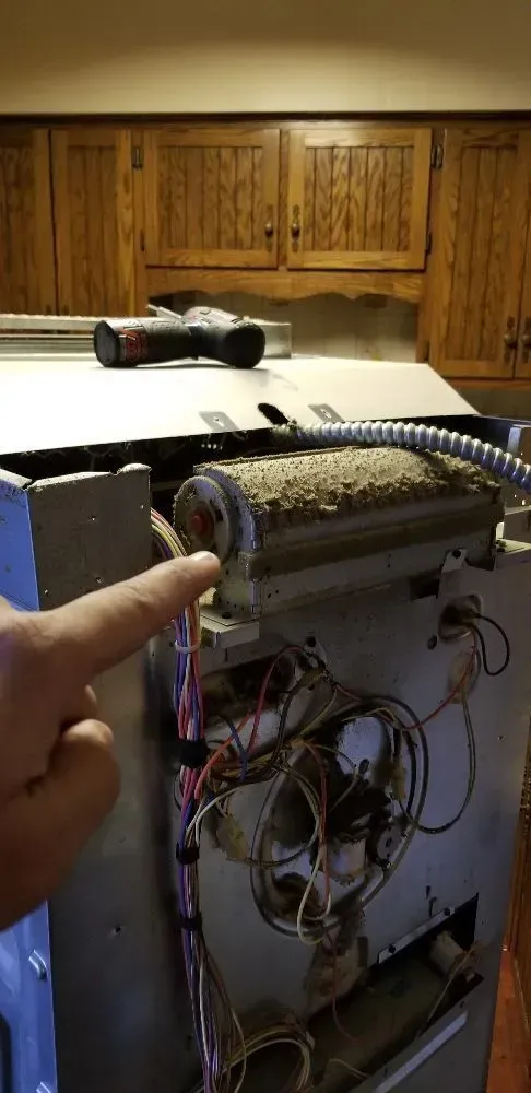 Person pointing at a dusty furnace component, likely a blower wheel. Wires and a power tool are visible.