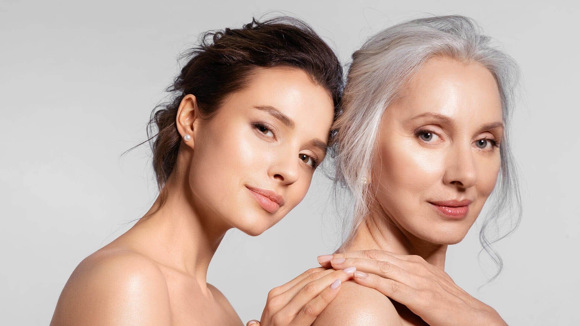 Two individuals with contrasting hair colors, dark and light, posing side-by-side with a calm expression against a gray background.