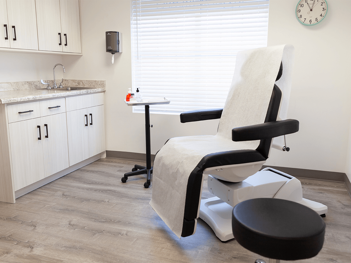 An examination room with a white adjustable chair, a small rolling side table, cabinetry with a sink, and a wall clock.