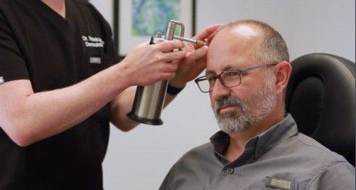 Technician sprays a man's scalp with a can while he sits in a salon chair, looking down.