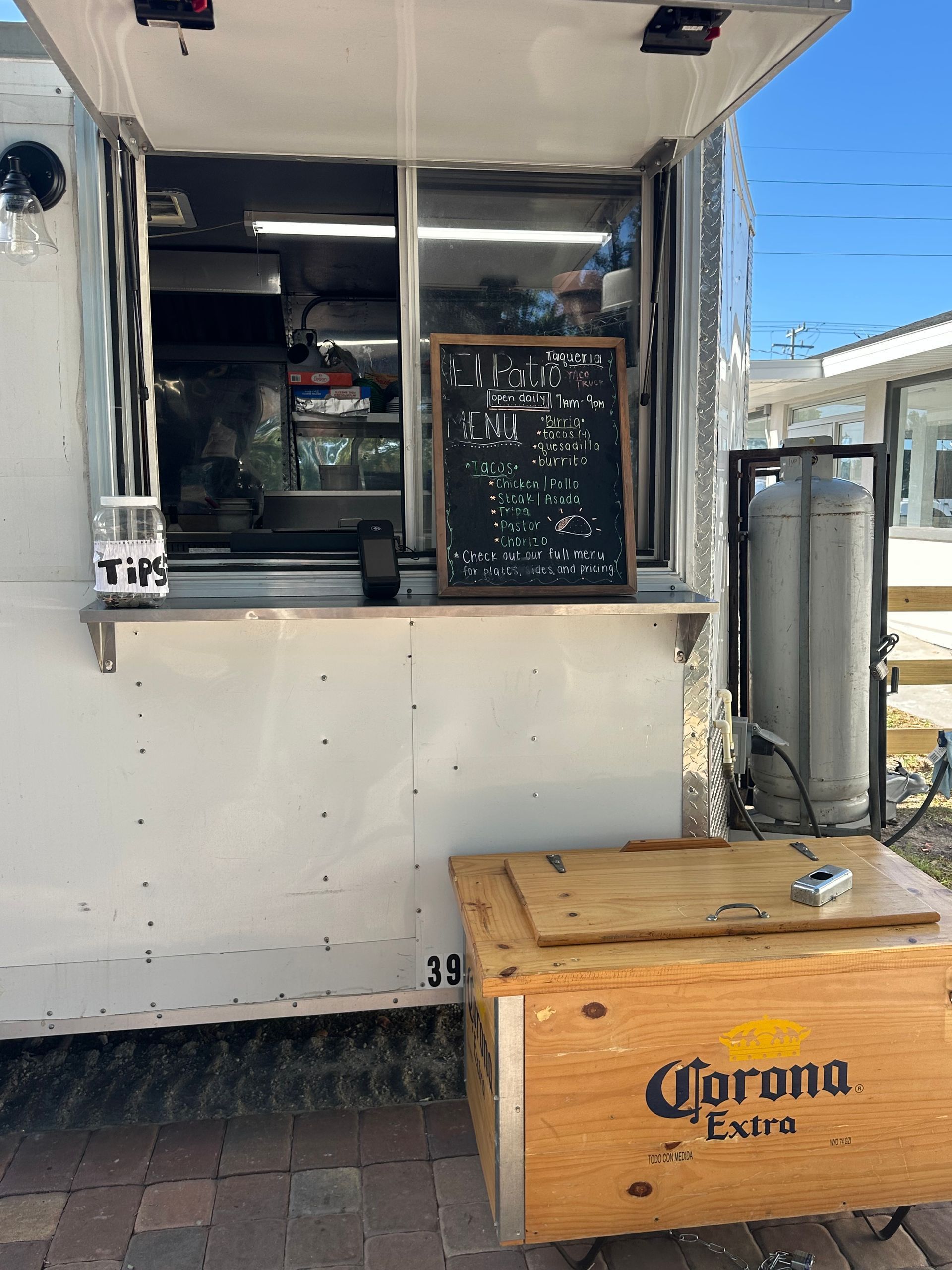 Food truck with open service window, chalkboard menu, and wooden Corona-branded box outdoors.