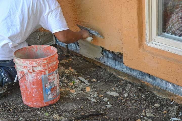 Worker scraping and patching damaged wall near a doorway, with an orange bucket on the ground
