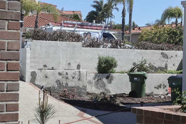 Residential driveway beside a white retaining wall, with a green trash bin and trees in the background
