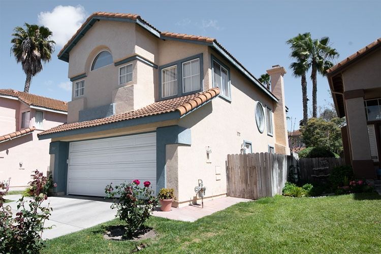 Two-story beige suburban house with garage, red-tiled roof, and a small front lawn under a clear sky