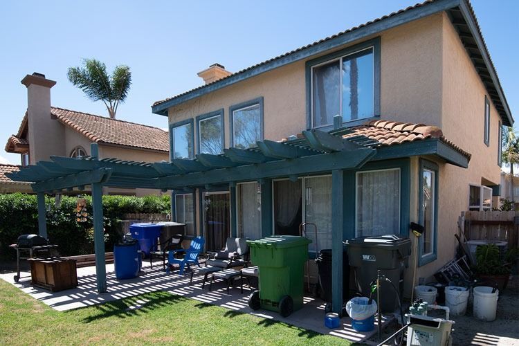 Two-story beige house with a covered patio, blue chairs, green bin, and a grassy backyard in sunlight