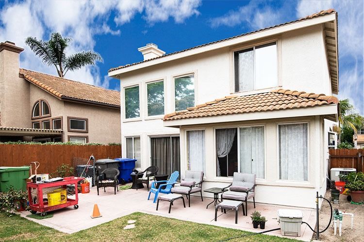 Backyard patio behind a two-story beige house with outdoor chairs, grill, and play equipment under a blue sky
