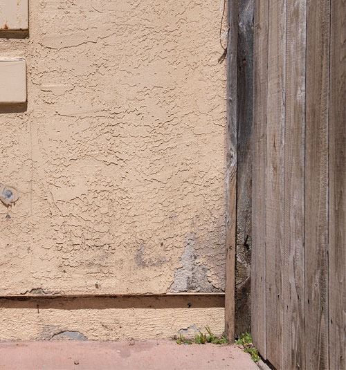 Beige stucco wall beside a weathered wooden fence, with a small gap at the bottom.