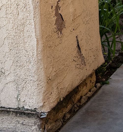Damaged stucco wall corner with peeling paint and exposed base near a concrete walkway
