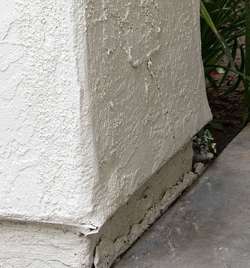 Cracked white stucco wall corner beside a concrete walkway and plants