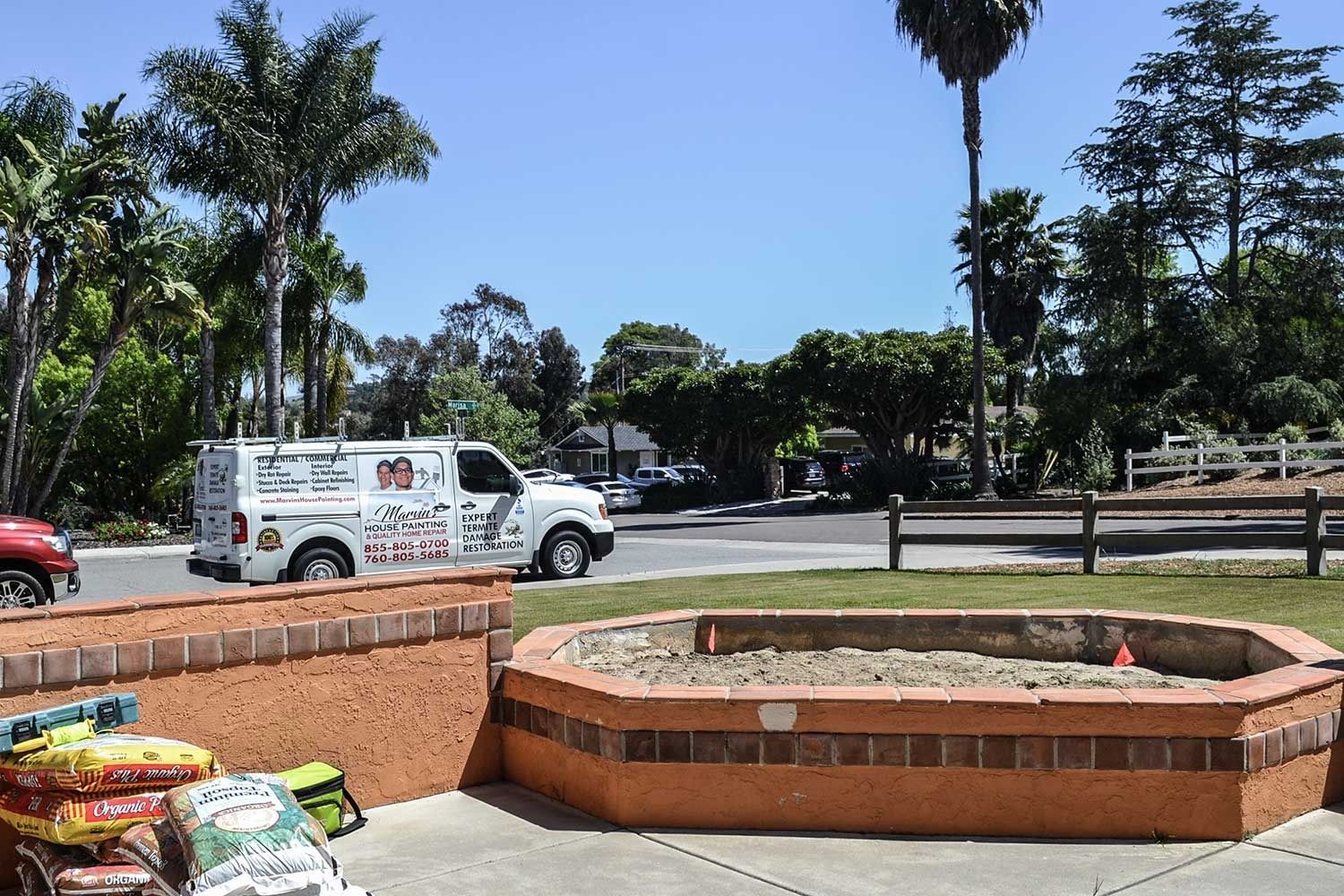 White truck parked by a brick planter in a sunny park with palm trees and a road in the background