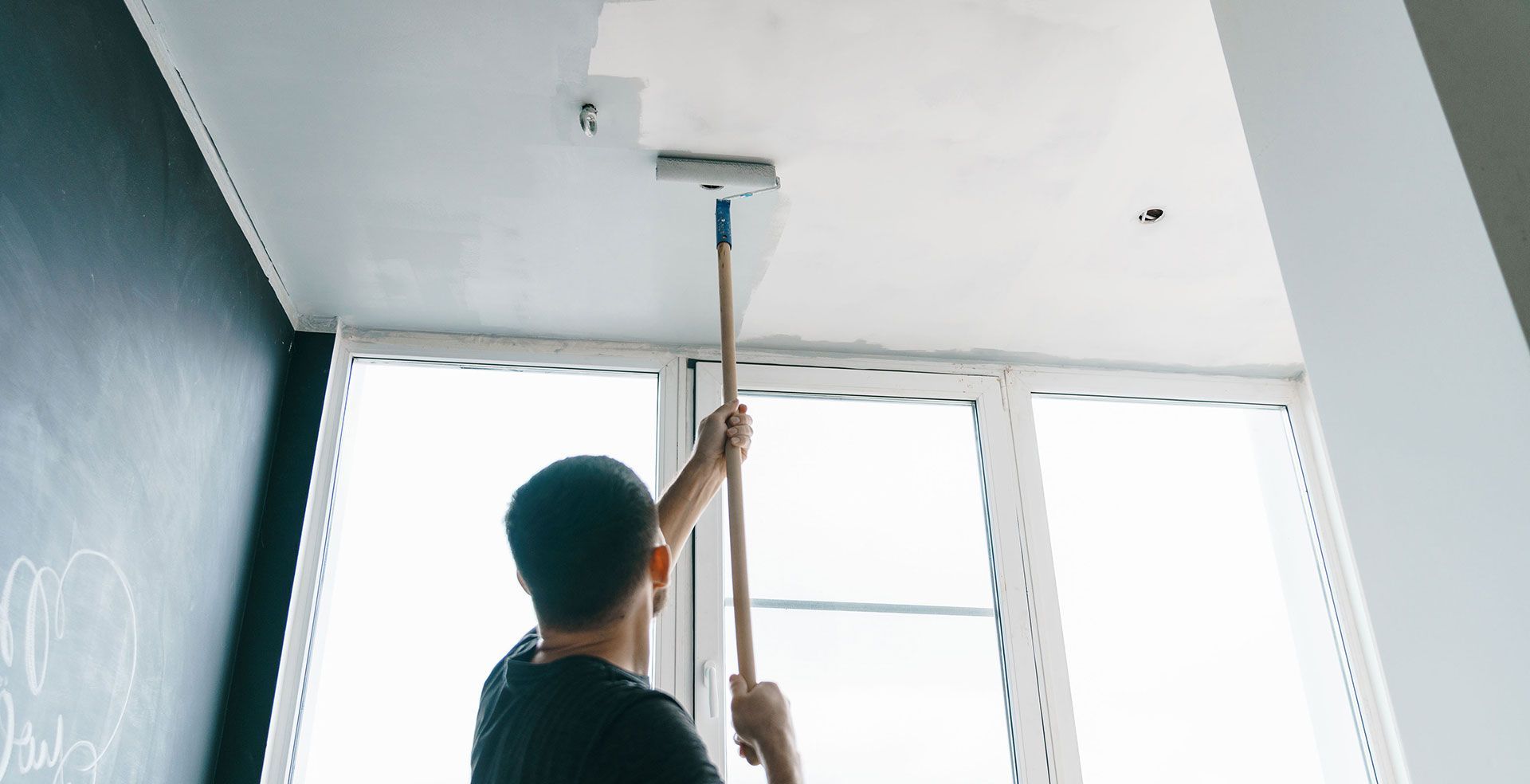 Person using a mop to clean a ceiling in a bright room with large windows