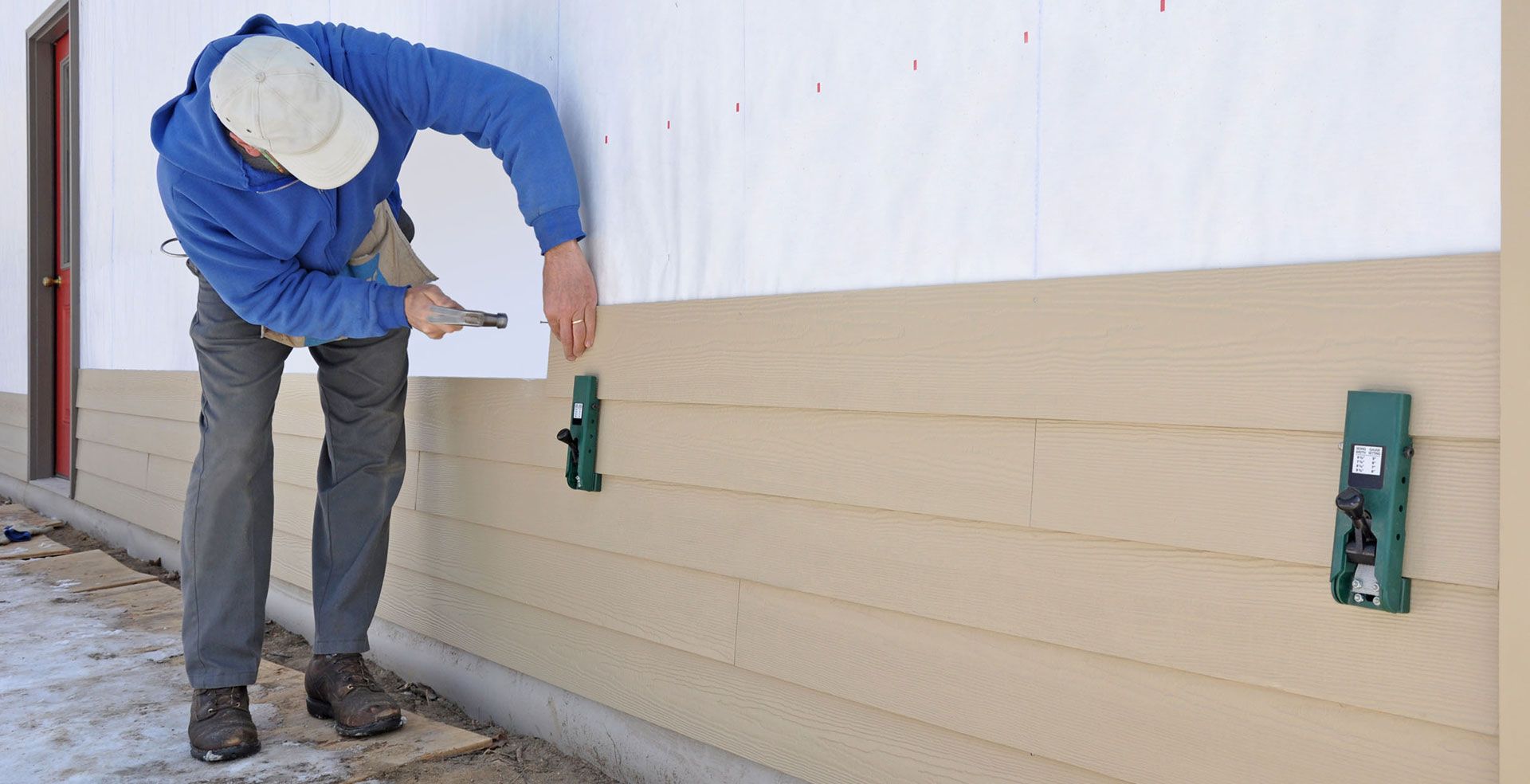 Worker in blue jacket painting beige exterior wall with a brush roller outdoors