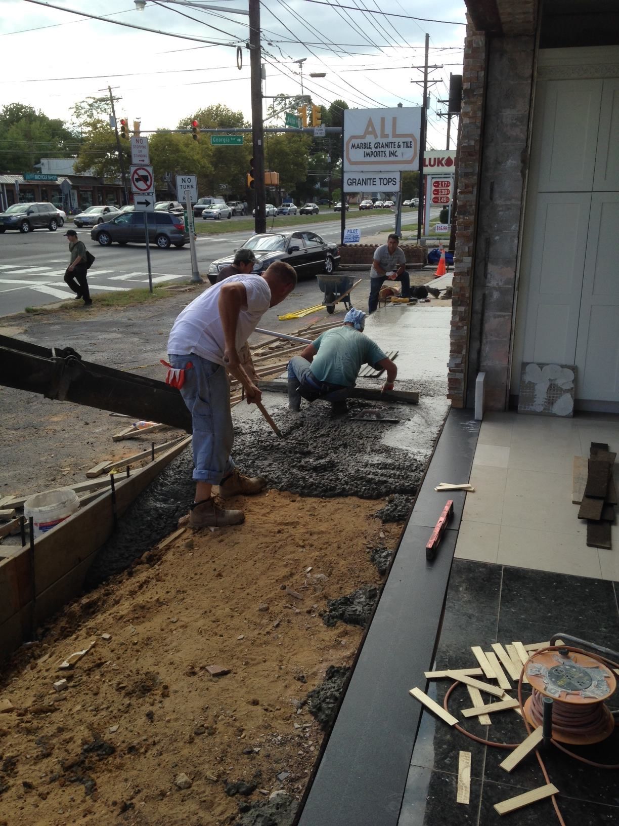 Two men are working on a sidewalk in front of a building with a sign that says all
