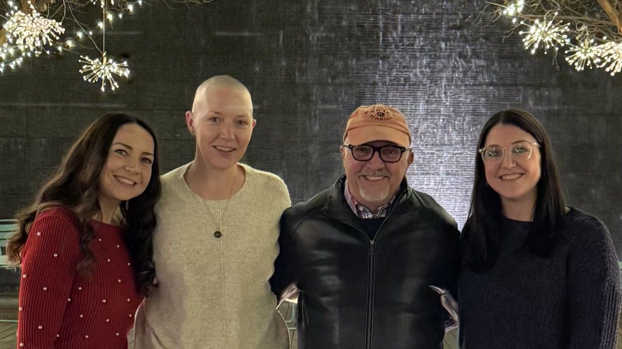 Four people smiling together in front of a water feature, string lights.