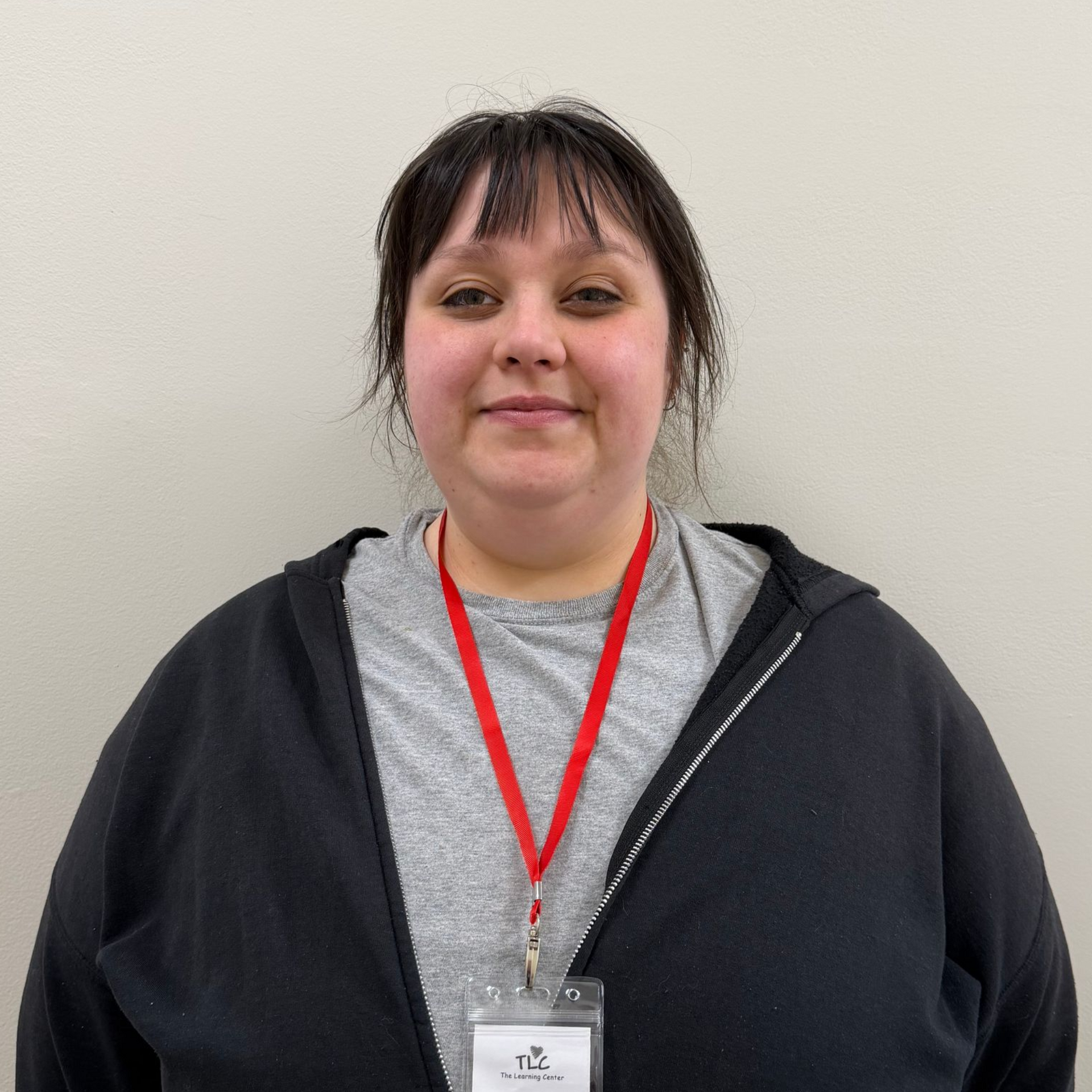 Woman wearing a lanyard and zipped black hoodie, standing in front of a neutral-colored wall next to a handicap restroom sign.