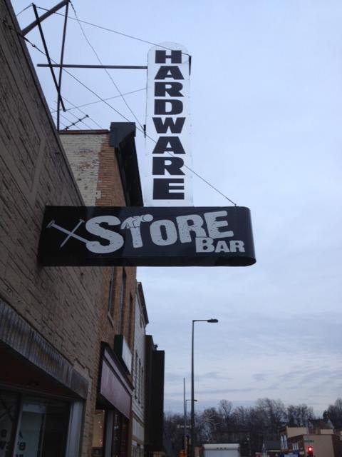 A hardware store bar sign hangs from the side of a building