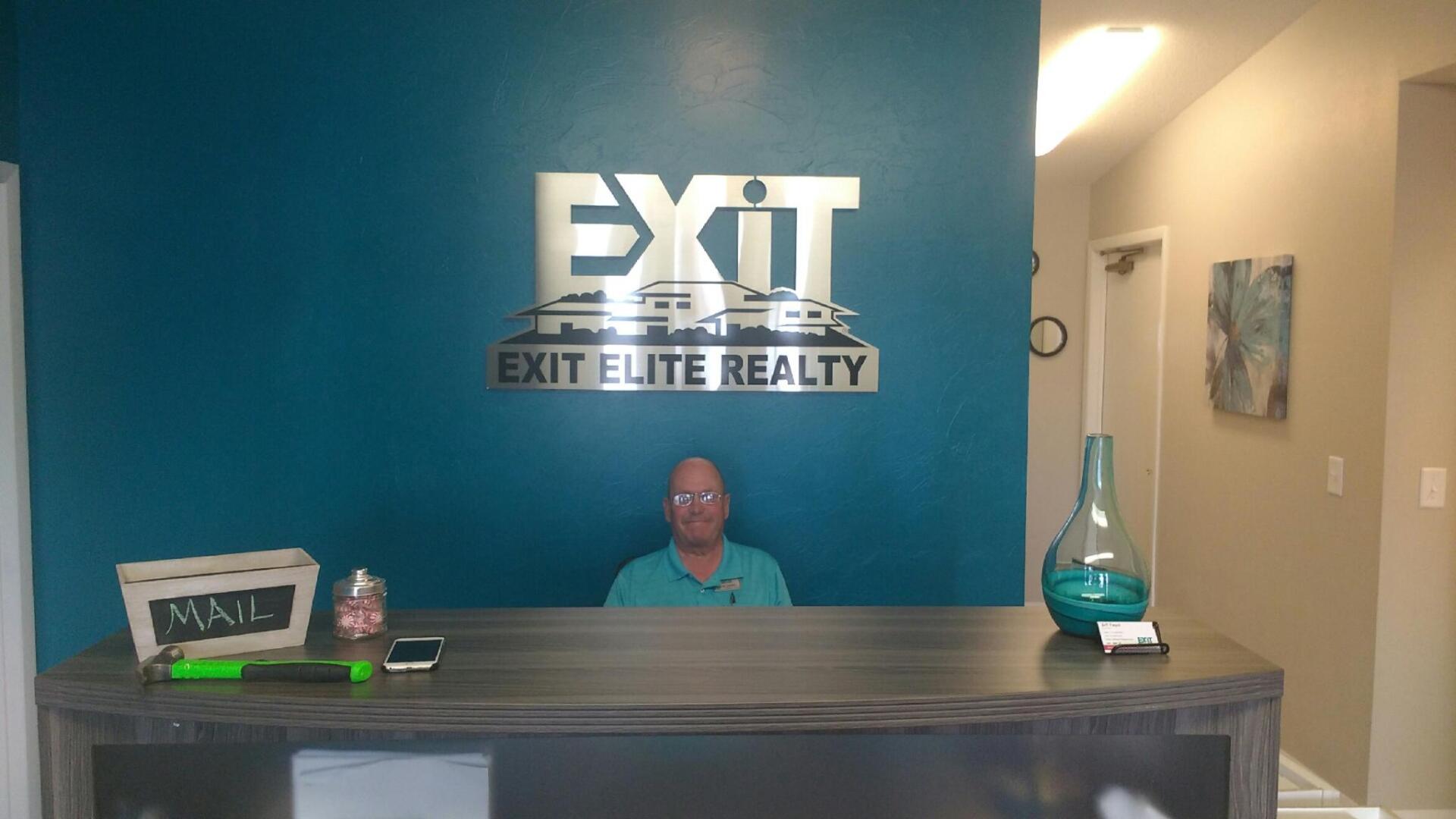 A man is sitting at a desk in front of a sign that says exit elite realty