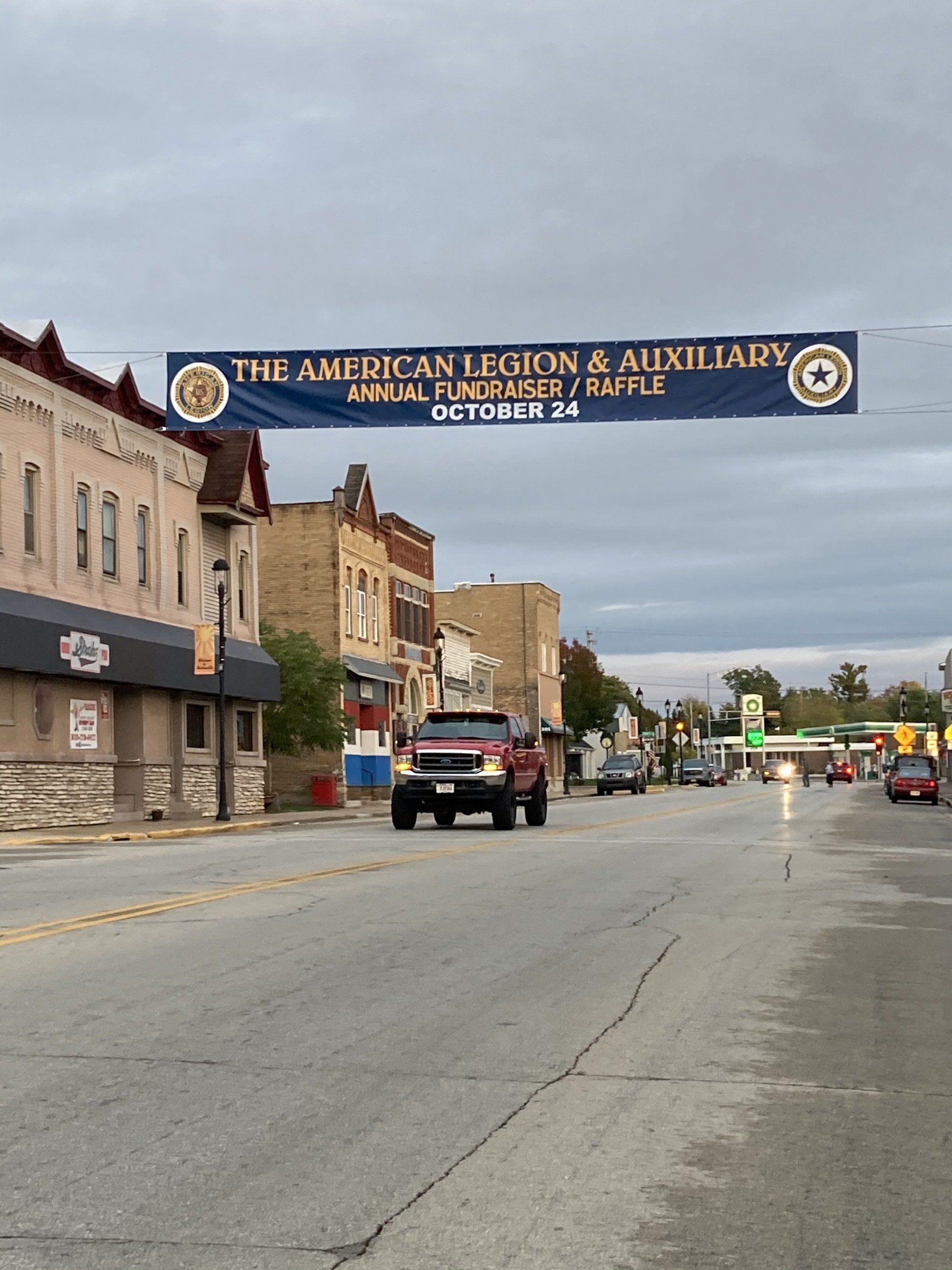 A red truck is driving down a street under a banner that says the american legion auxiliary