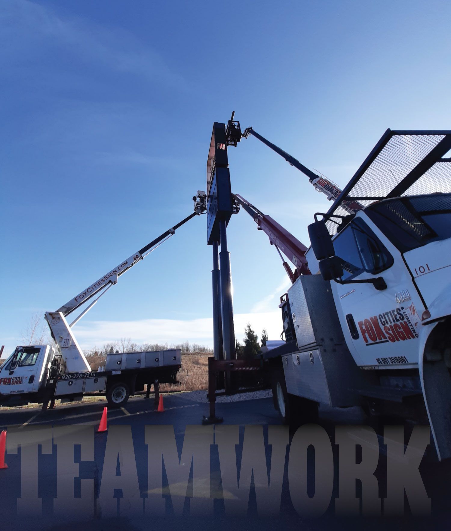 Crew using cranes to raise a large 'TEAMWORK' sign on a sunny outdoor worksite.