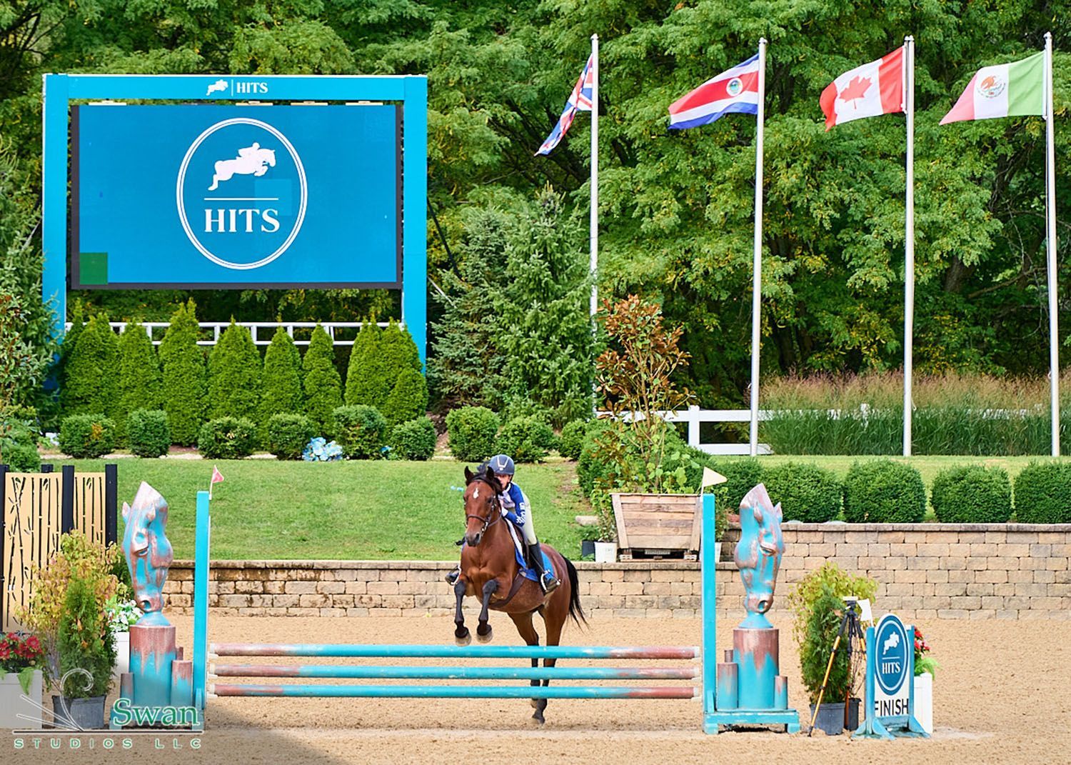 Horse and rider clearing a jump at an outdoor equestrian arena with HITS sign and flags.