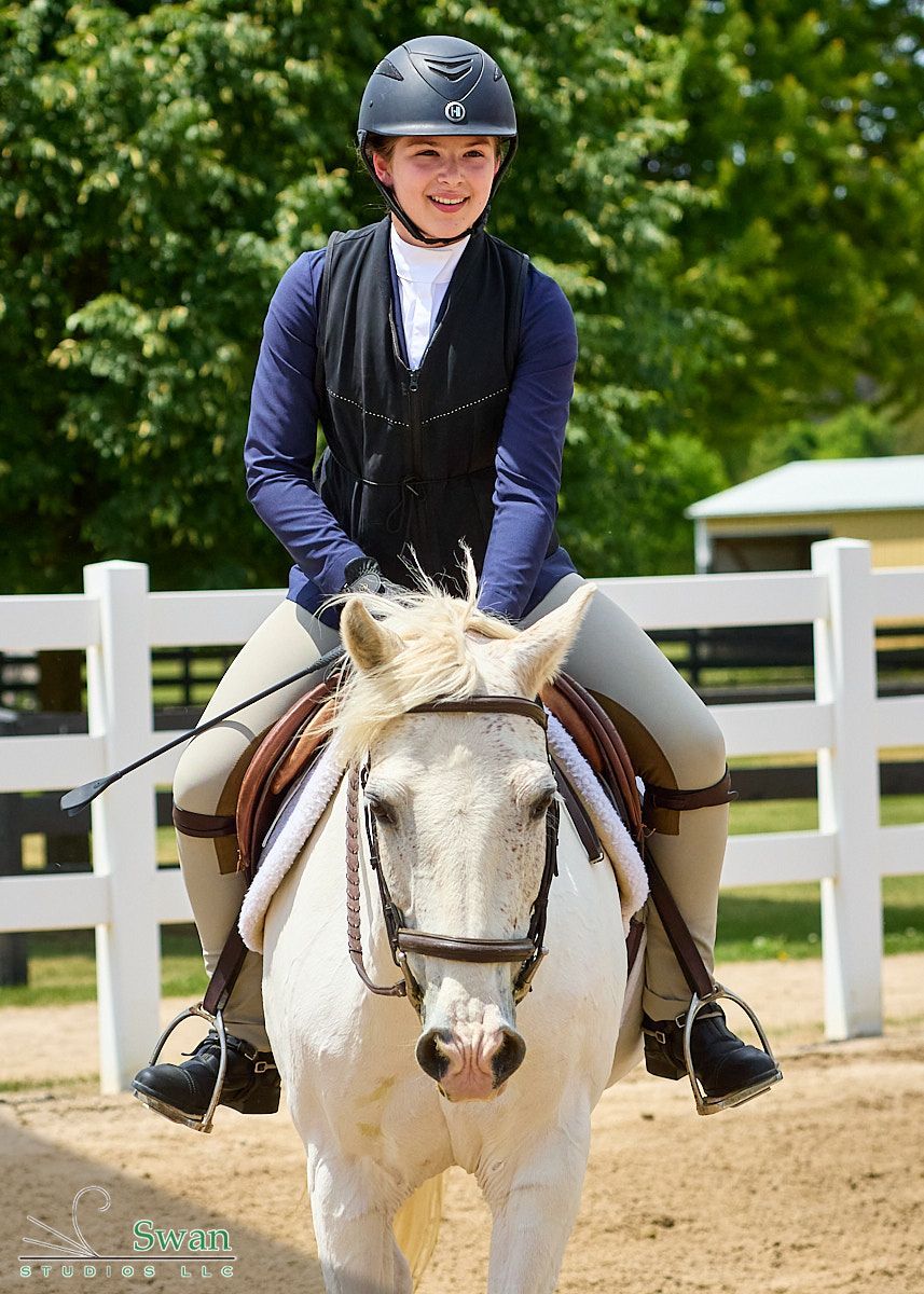 Rider in helmet on white horse in an outdoor arena, with a white fence and green trees behind.