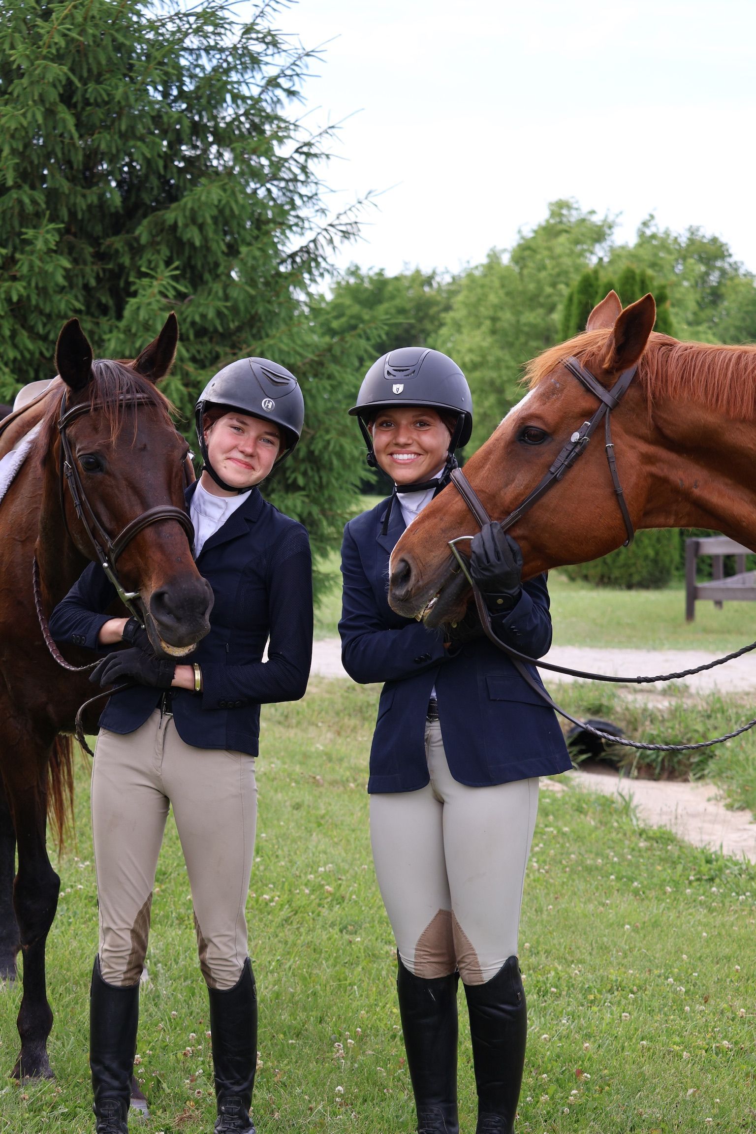 Two helmeted riders in navy show jackets stand with two horses in a grassy outdoor setting.