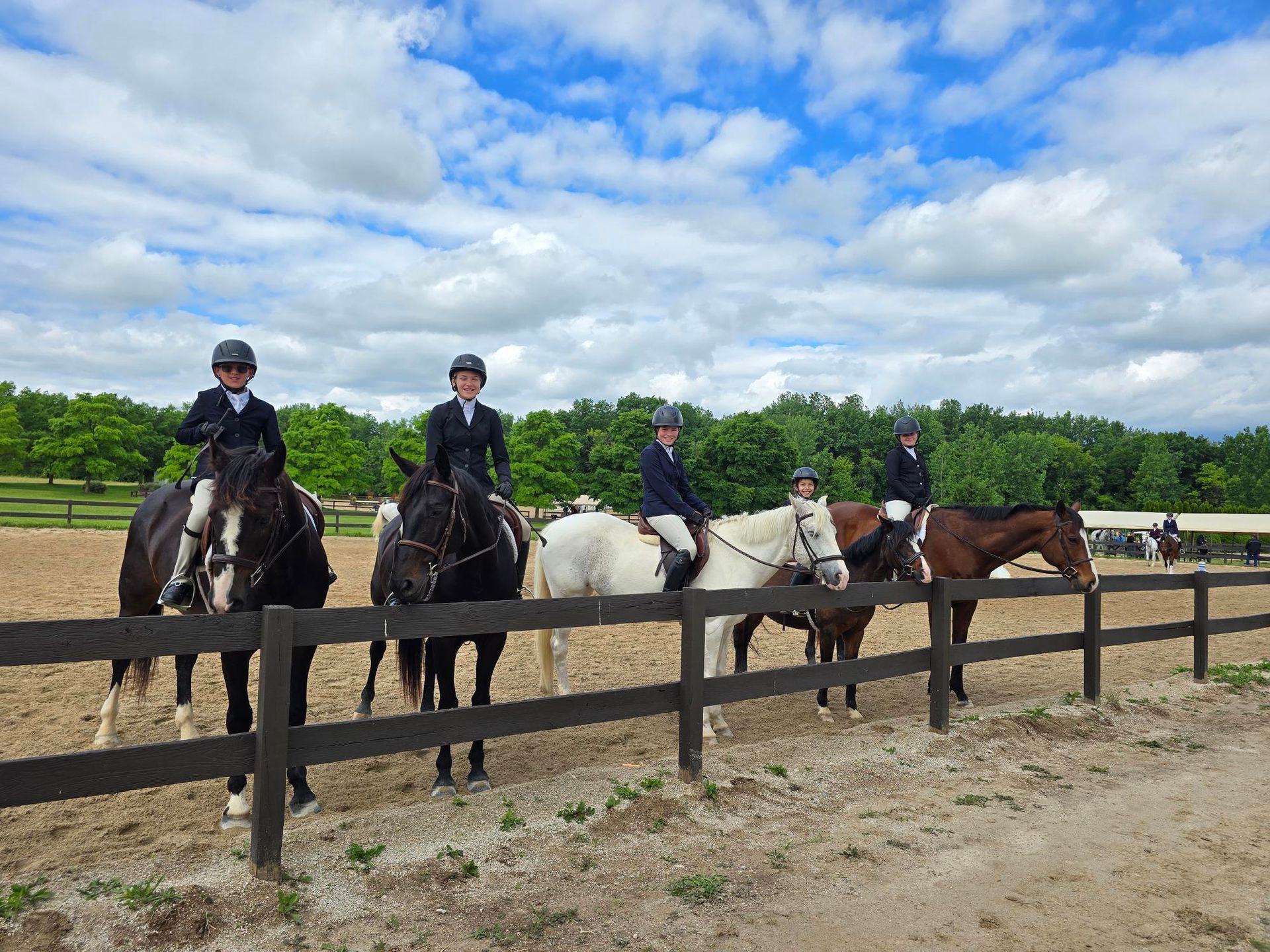 Five horseback riders lined up by a fence in a grassy field under a cloudy sky.