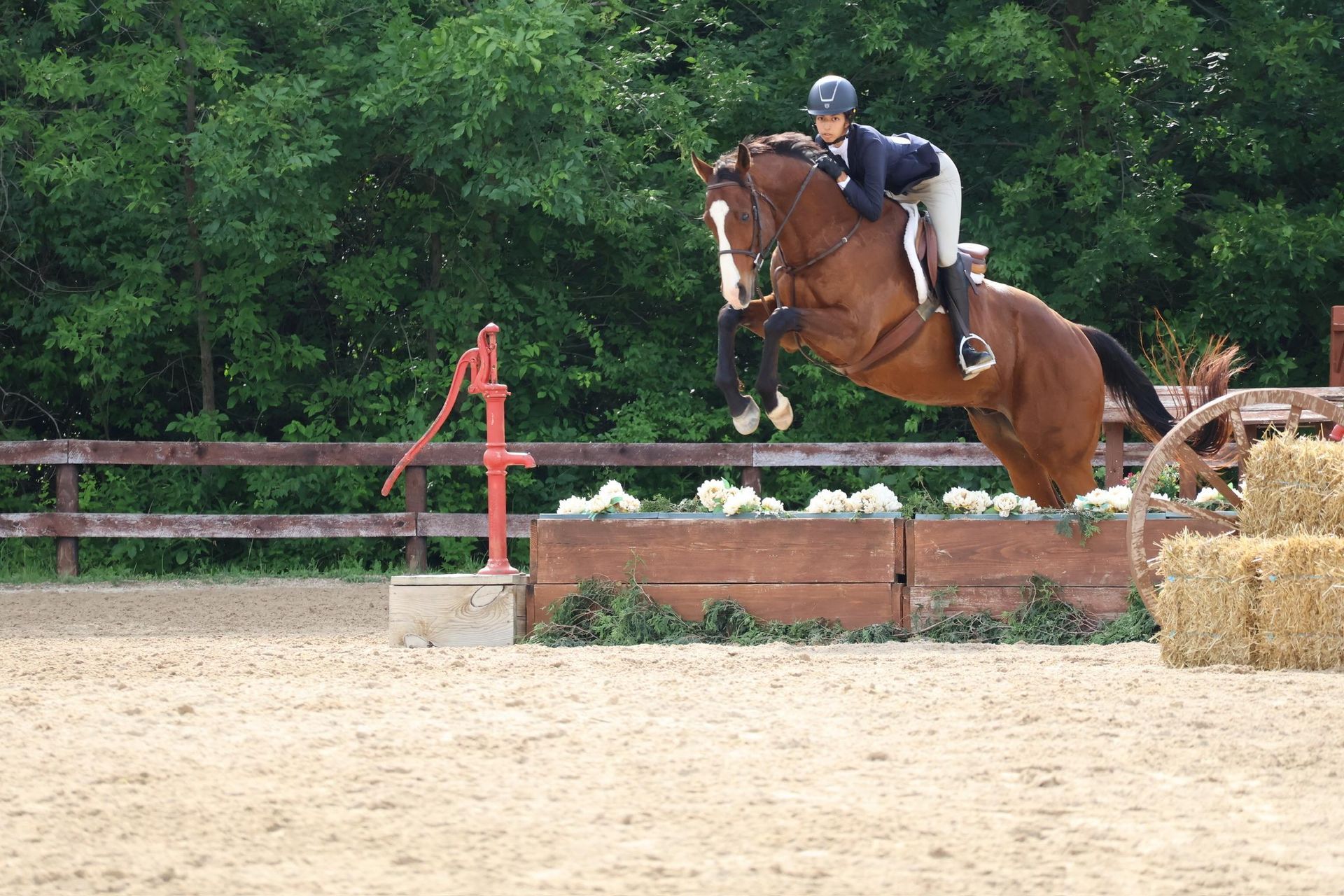 Rider and brown horse clearing a jump in an outdoor arena, with hay bales and a wooden fence behind.