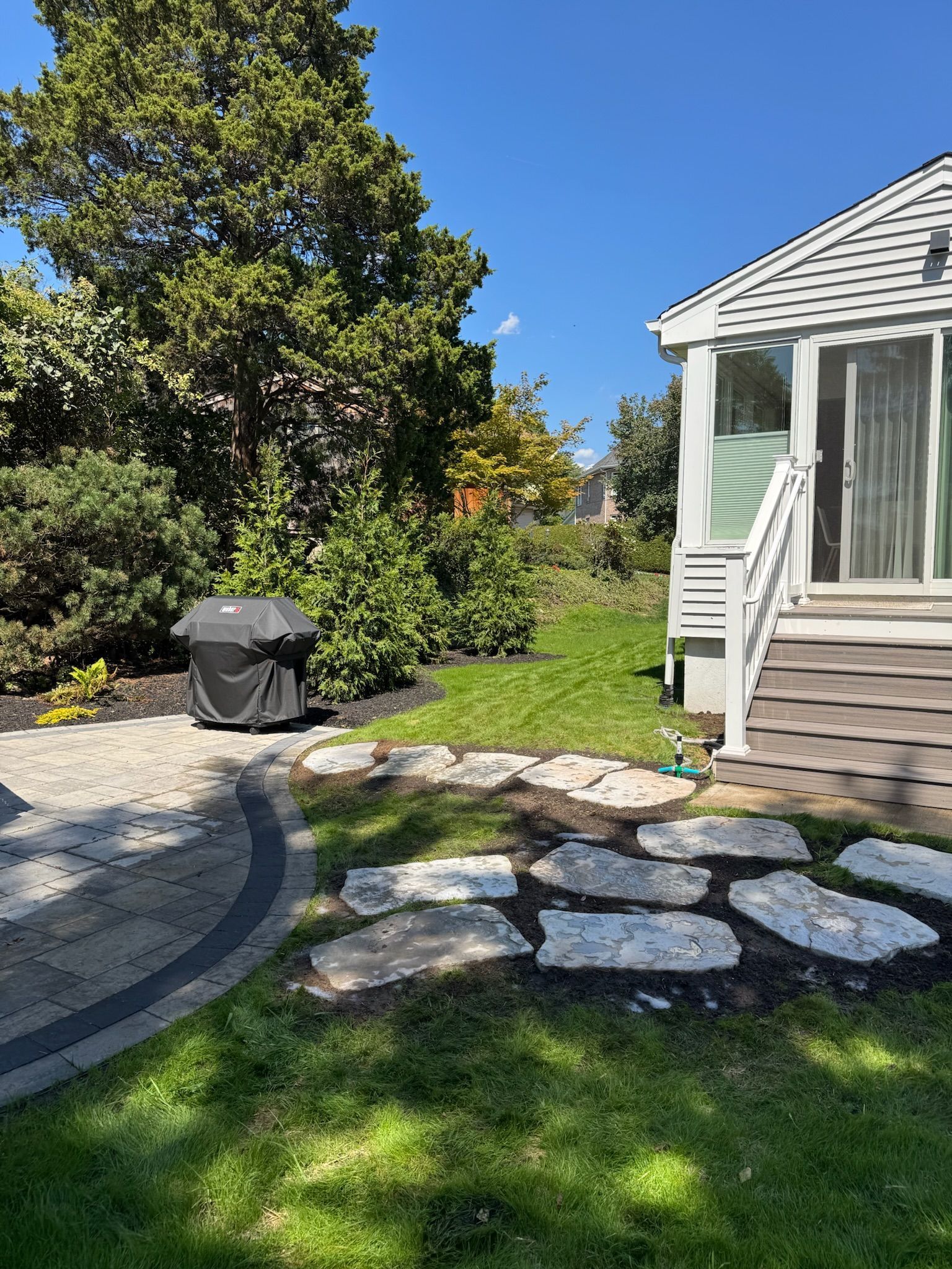 Patio with grill, stone path, grass, and white house with stairs. Sunny day.