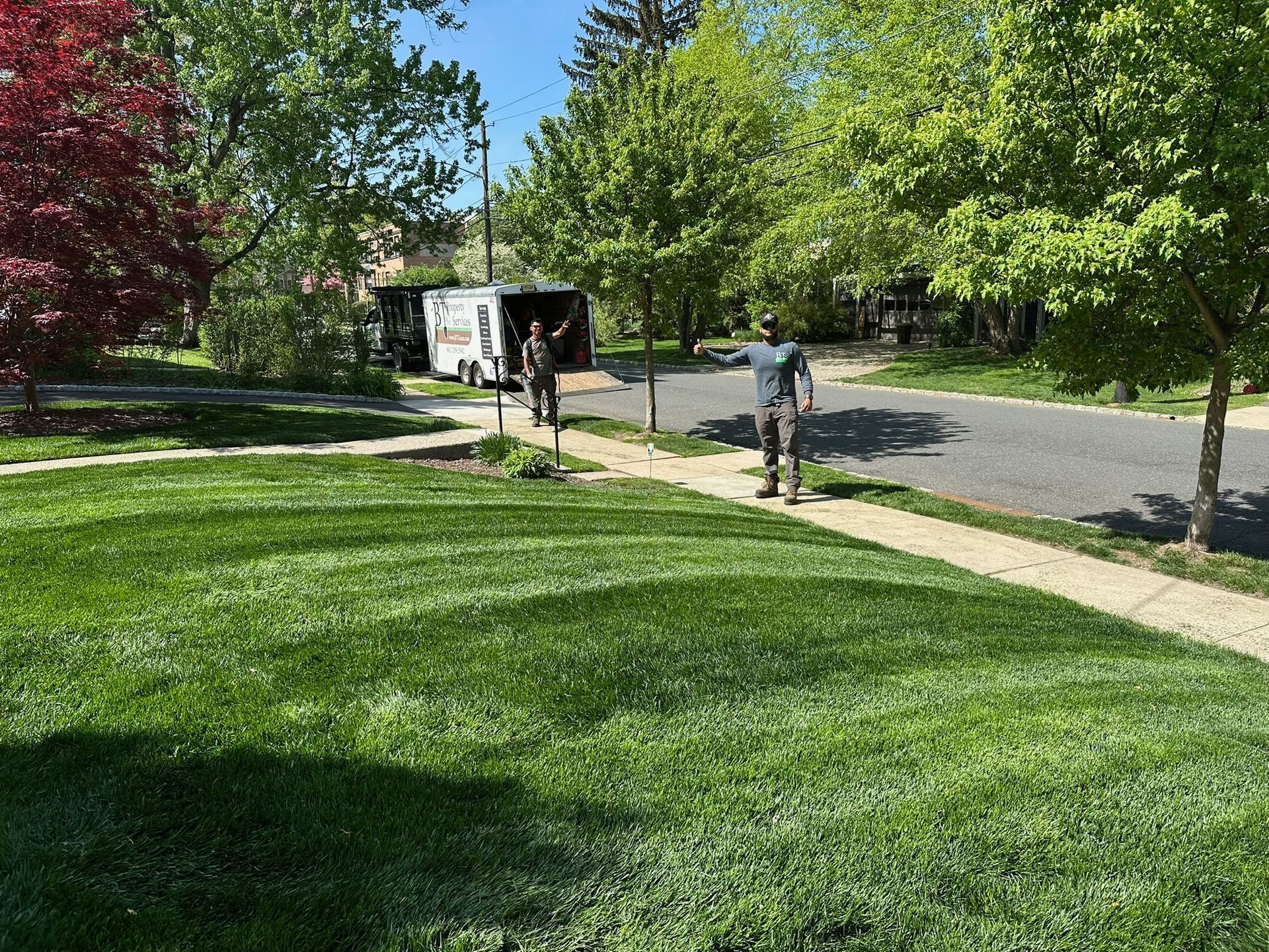 Lawn with two people by a trailer on a street. Green grass, trees, and blue sky.